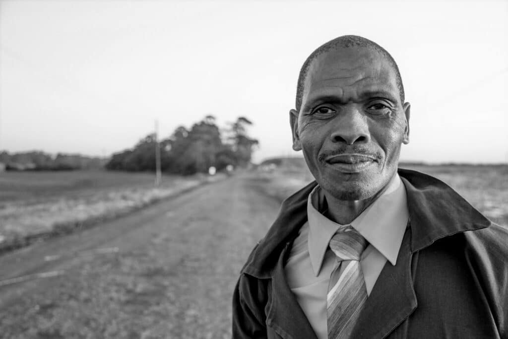 A man wearing a coat and tie standing on a rural road, with fields and trees in the background.