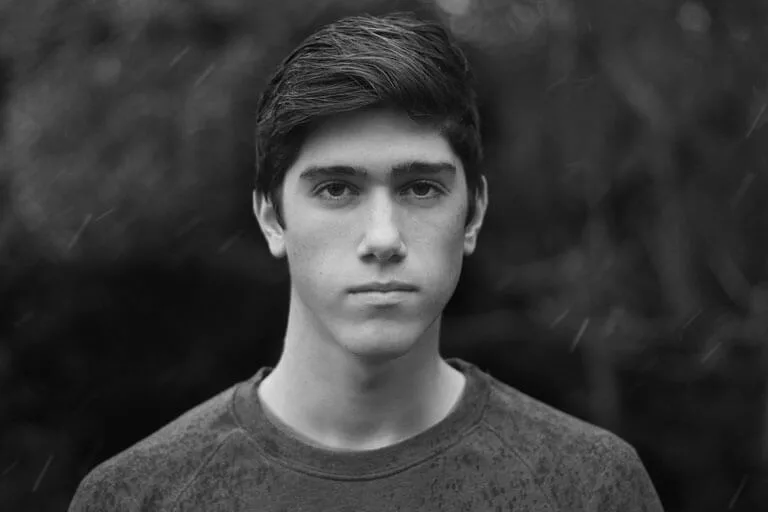 Black and white portrait of a young man with short dark hair, looking directly at the camera with a neutral expression, blurred outdoor background.