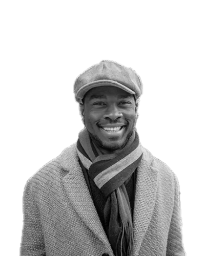 Black and white portrait of a smiling man wearing a flat cap, a warm scarf, and a textured coat, standing against a light background.