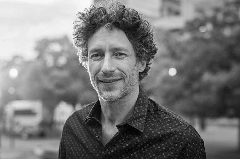 Black and white photo of a man with curly hair and a patterned shirt, smiling slightly while standing outdoors with trees and soft lights in the background.